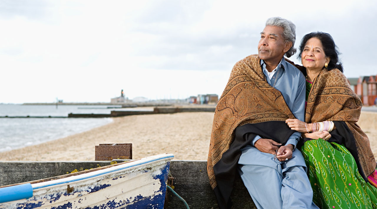An older Indian couple sit on the beach, wrapped in a blanket staring off into the distance.