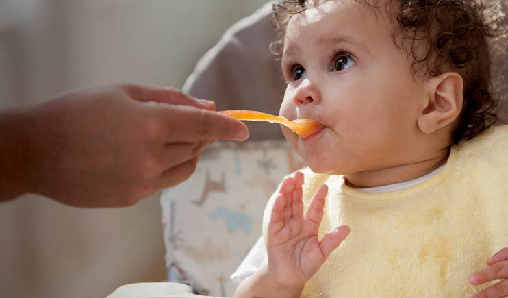 A person's hand spoon-feeding a baby in a yellow bib.