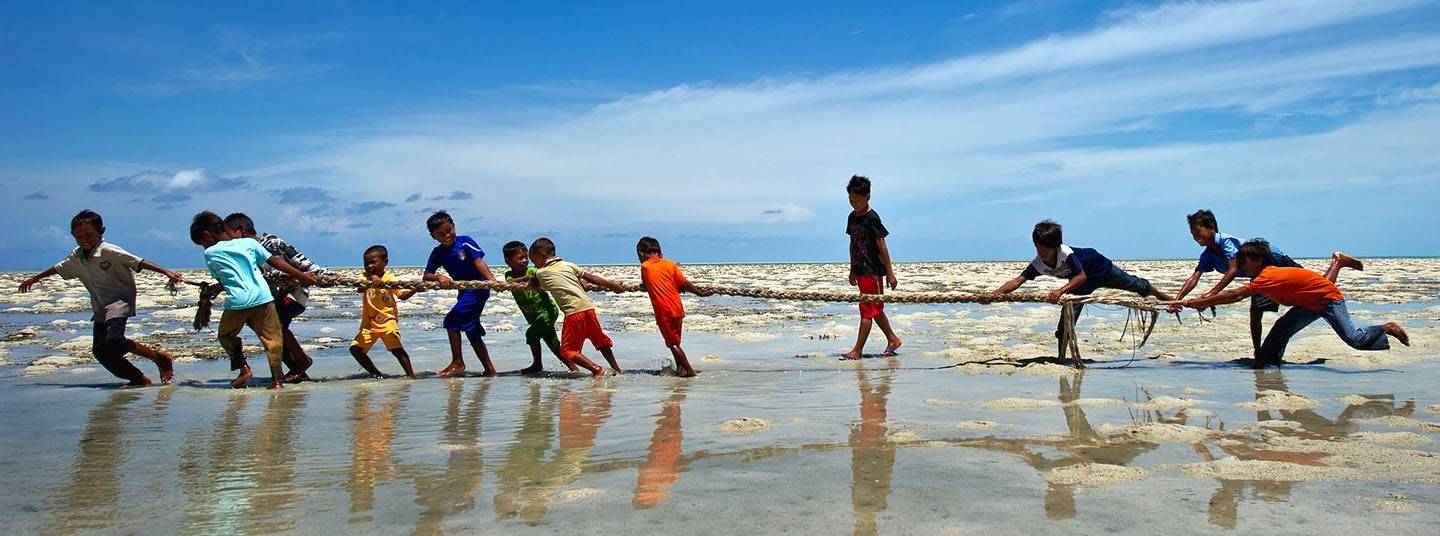 Under a blue sky, young Indian boys in bright clothing play tug of war in shallow water.