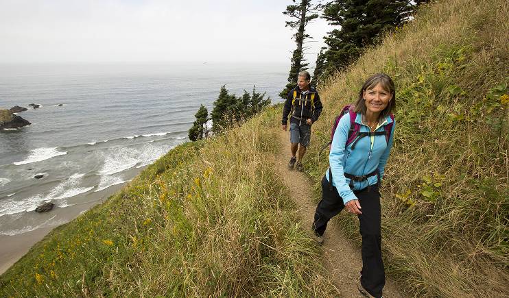 A middle-aged couple hikes along a narrow, grassy path on a hillside above the ocean. Both wear backpacks and outdoor clothing. 