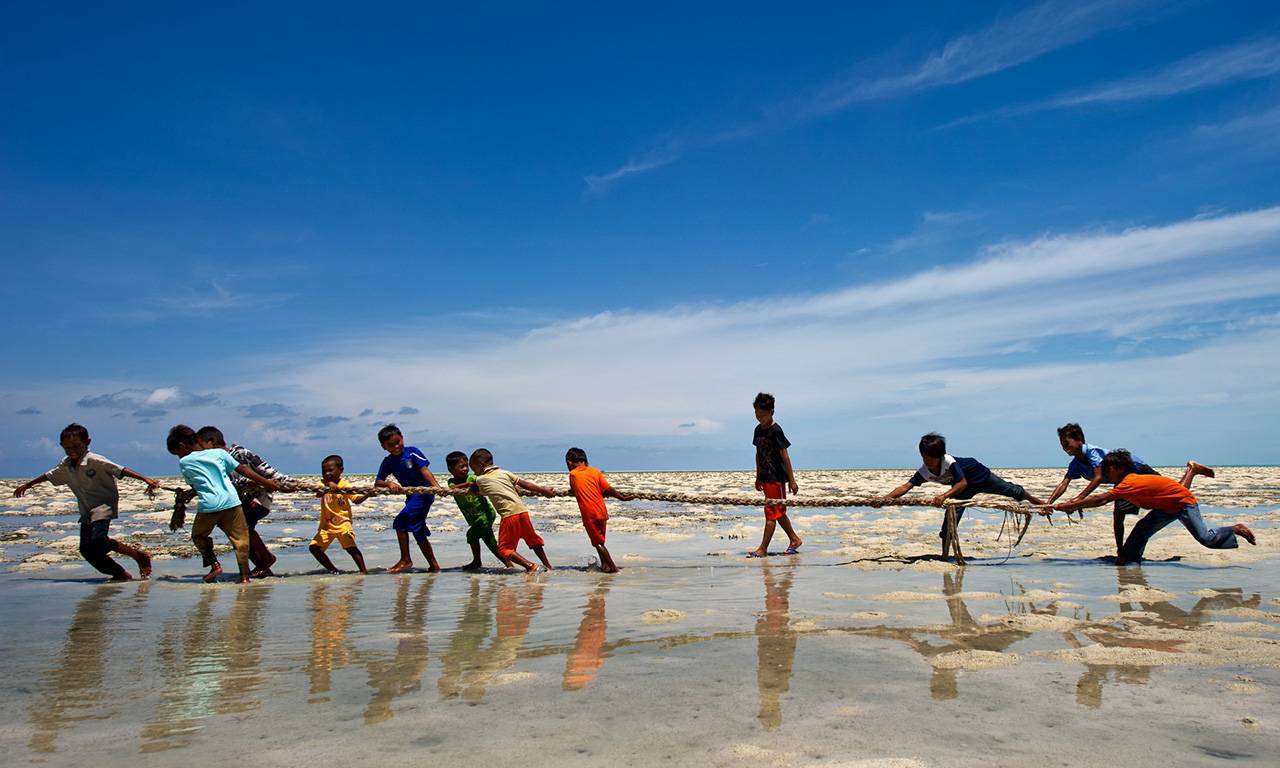 Under a blue sky, young Indian boys in bright clothing play tug of war in shallow water.