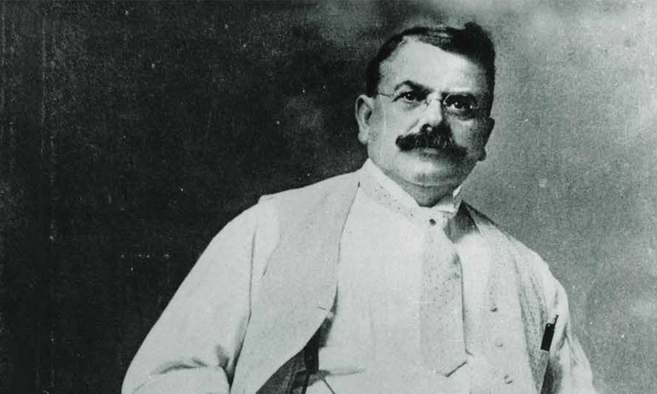 Photo en noir et blanc de Wallace C. Abbott.  Il a des cheveux noirs courts, une grosse moustache foncée et porte des lunettes, une chemise blanche, une veste de costume et une cravate.